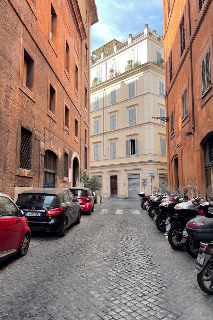 Explore a picturesque Roman street lined with cars and motorbikes amidst classic architecture.