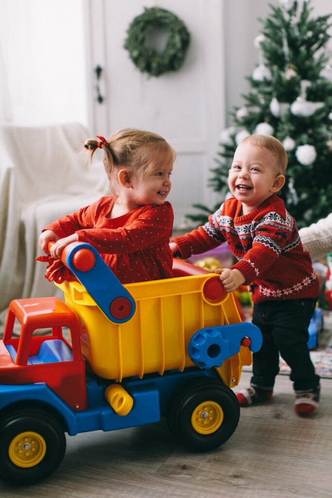 Two toddlers happily play with a toy truck in a festive, indoor setting with a Christmas tree.